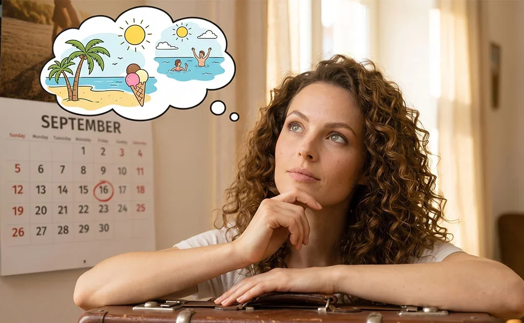 Una donna con capelli ricci appoggiata a una valigia guarda pensosa un calendario a muro sulla pagina di settembre con la data del 16 cerchiata, mentre immagina le vacanze estive al mare.